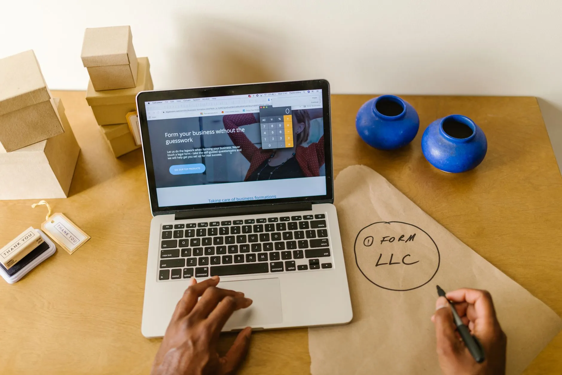 A small business owner at a laptop with a calculator open, shipping boxes on the desk, writing "form LLC" on kraft paper
